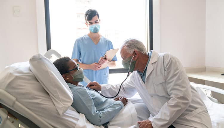Senior male doctor checking black hospitalized female patient listening to her heart and nurse standing next to them all wearing protective facemasks